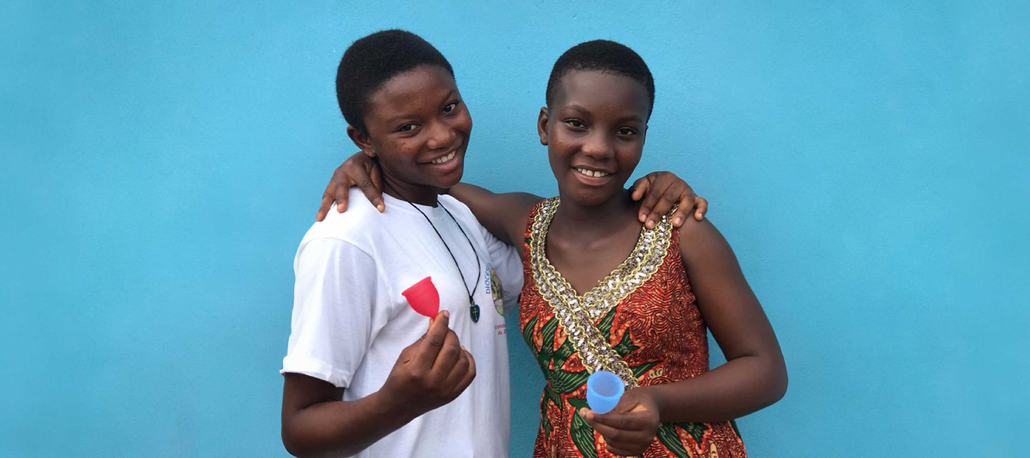 Two smiling girls holding menstrual cups while standing together against a blue background.