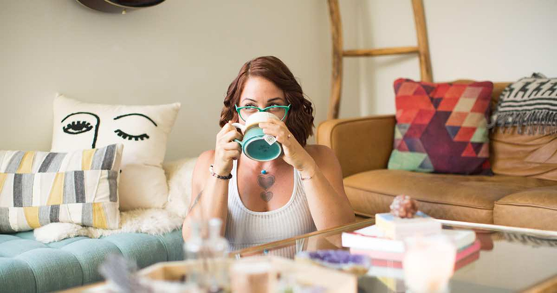 Woman drinking from a mug while sitting in a cozy living room with colorful pillows.