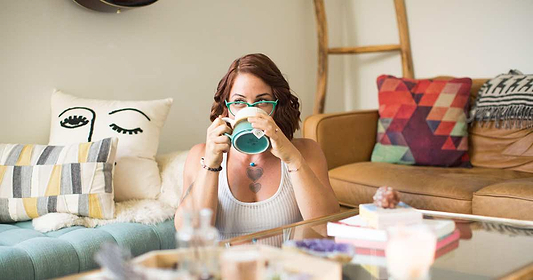 Woman drinking from a mug while sitting in a cozy living room with colorful pillows.