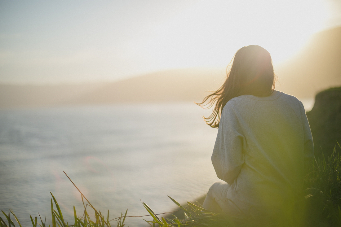 Person sitting on a grassy cliff overlooking the ocean at sunset, with sunlight glowing behind them.