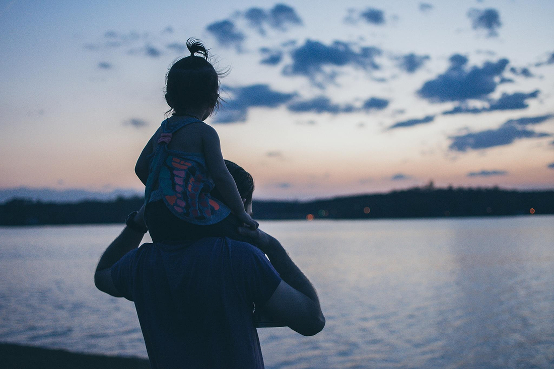 Parent carrying a child on their shoulders by a lakeside at dusk, watching the calm water under a cloudy evening sky.
