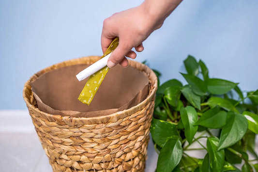 Hand placing a wrapped hygiene product into a woven trash bin beside a green houseplant.