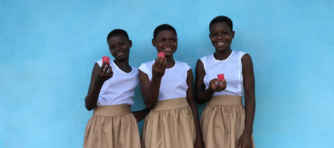 Three smiling girls holding reusable menstrual cups while standing against a blue wall.
