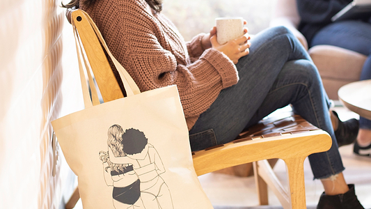Canvas tote with body-positive illustration beside person relaxing in chair with a mug.
