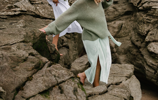 Person in oversized sweater and flowing skirt walking barefoot across rocky shoreline.