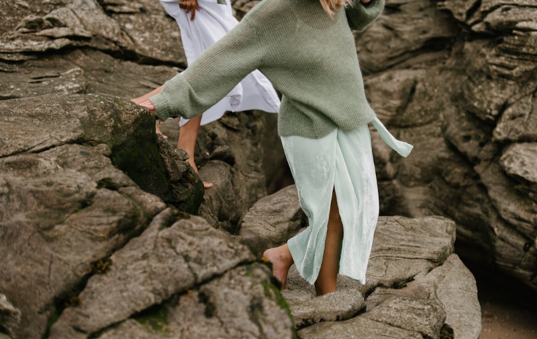 Two women carefully walking barefoot across rocky coastal terrain outdoors