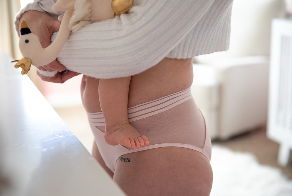 Person holding baby while wearing light pink mesh underwear indoors.