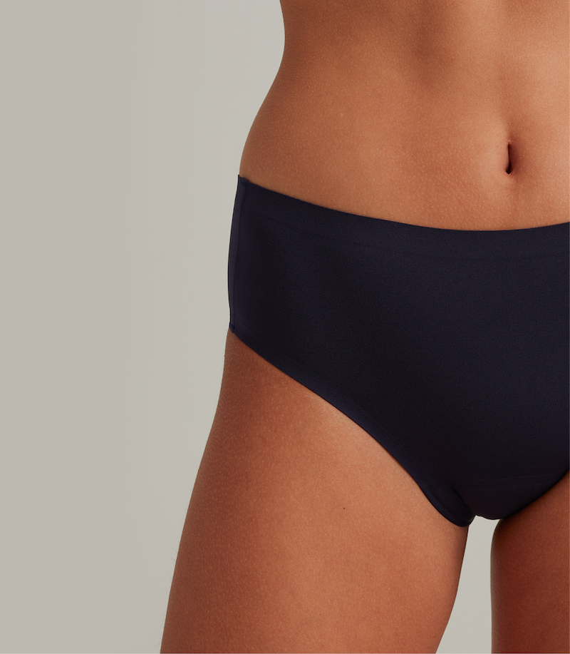 A close-up shot of a person's midsection and upper thigh modeling smooth, dark navy seamless underwear against a plain light gray background..