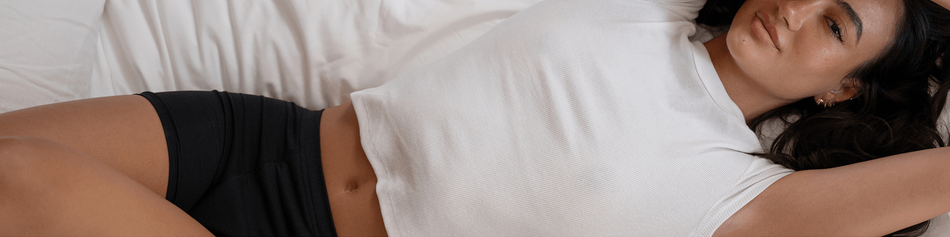 A model in Saalt's new Cotton Sleep Short leakproof period underwear in color Volcanic Black, laying on a bed with a white comforter. The tone is confident and relaxed.