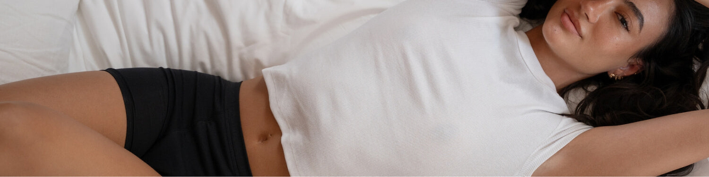 A model in Saalt's new Cotton Sleep Short leakproof period underwear in color Volcanic Black, laying on a bed with a white comforter. The tone is confident and relaxed.