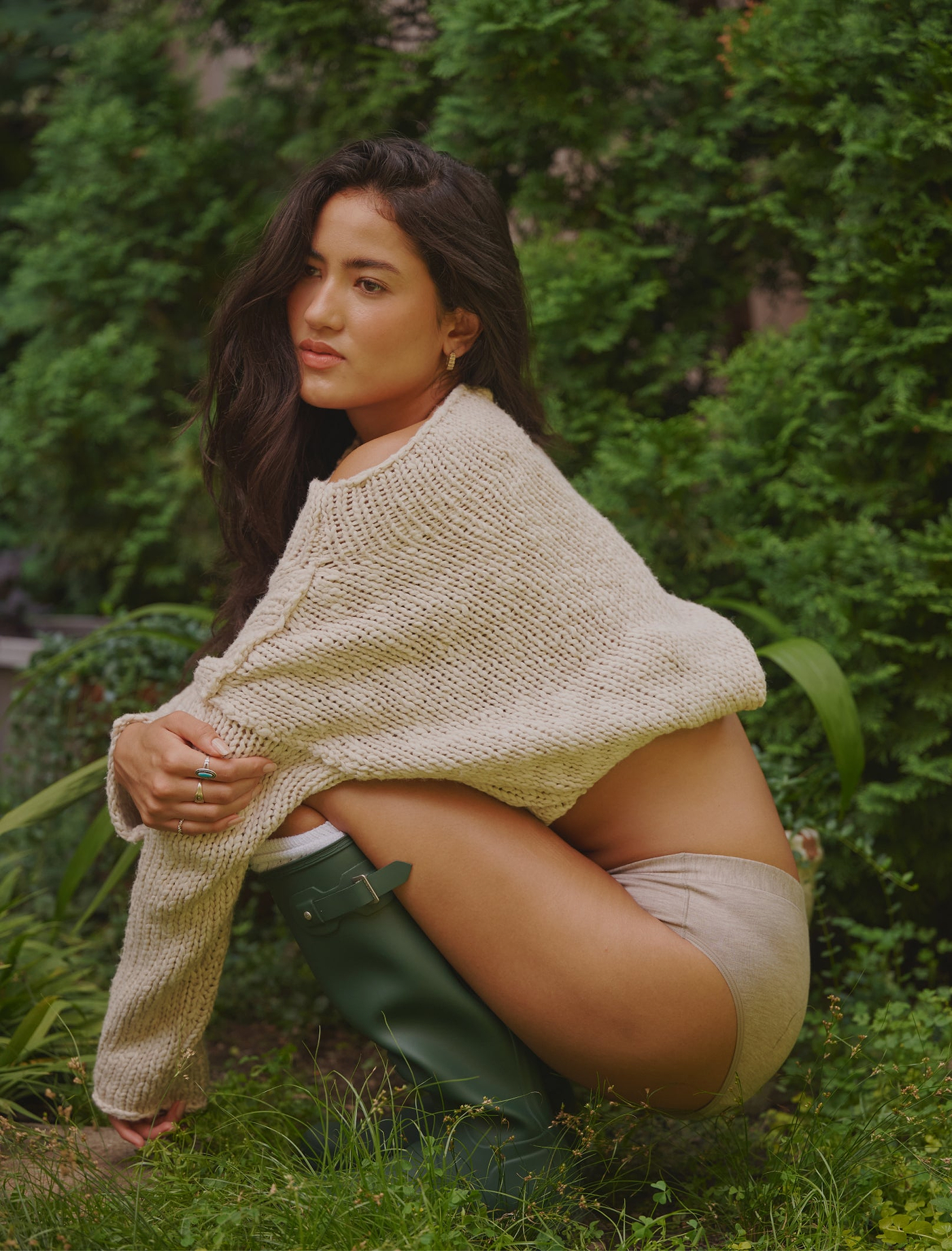 Model crouching in a garden wearing an oatmeal Leakproof Comfort brief, chunky knit sweater and green rain boots