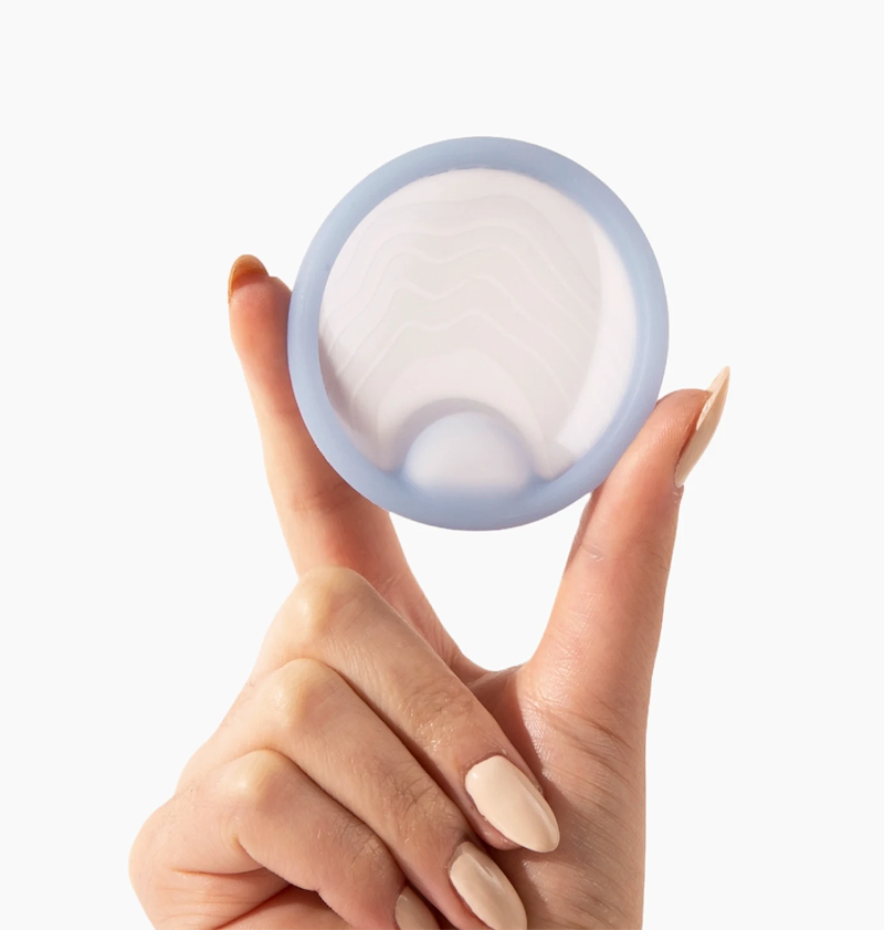 A close-up of a hand holding a light blue menstrual disc between two fingers against a plain white background. The disc features a firm, rim and a soft, wavy-patterned center with a molded finger notch for removal.