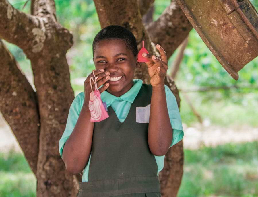 A smiling young girl in a school uniform holds up a red menstrual cup and its pink carrying pouch outdoors.
