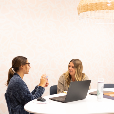 two females sitting at an office table talking to each other in front of a laptop