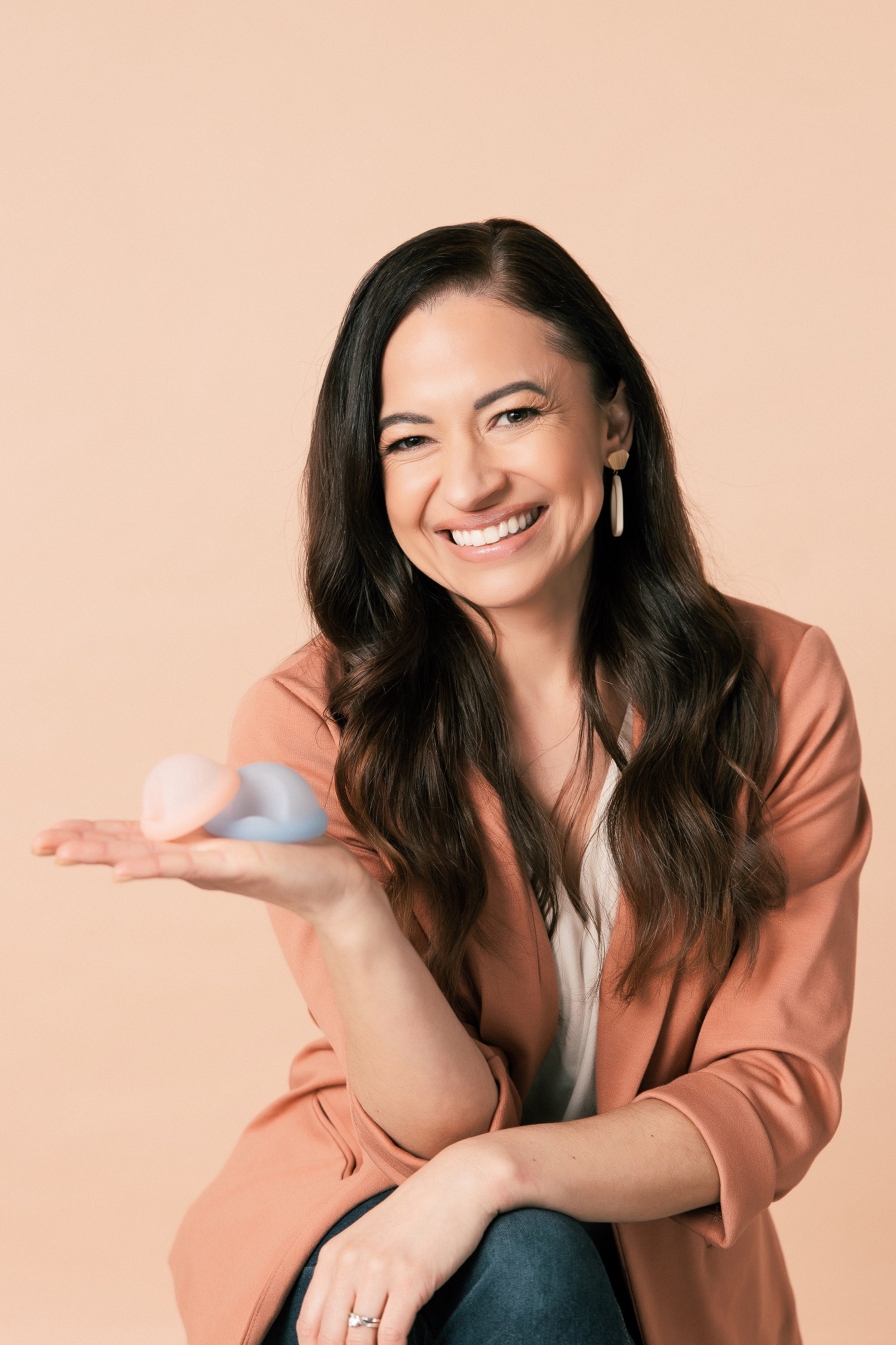 A smiling woman in a tan blazer holding a pink and a blue menstrual cup in her open palm.