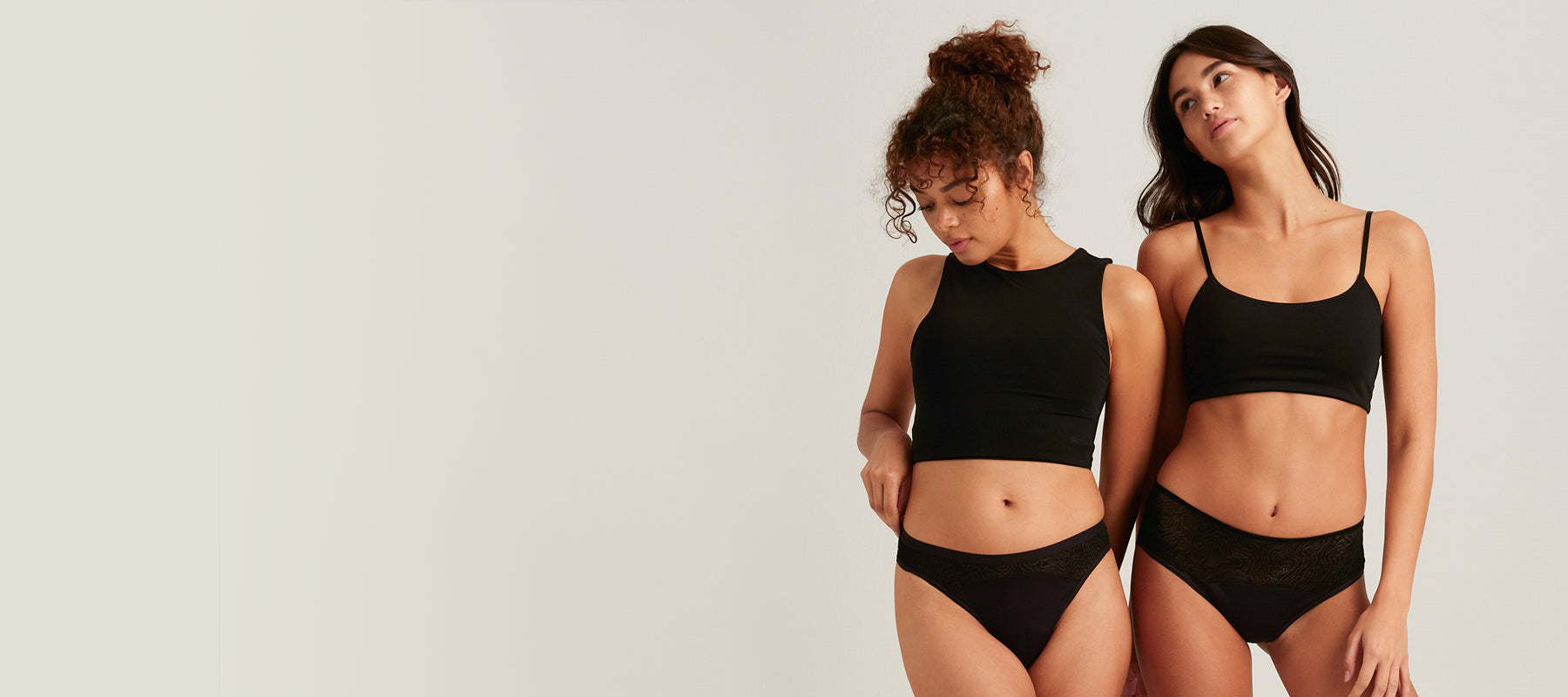 Two women wearing black period underwear and black bralettes from Saalt’s Lace Collection. The woman on the left wears a high-neck crop top with Saalt Leakproof Lace thong underwear, and the woman on the right wears a spaghetti strap bralette with Saalt Leakproof Lace Hipster underwear. Both pairs feature subtle lace detailing.