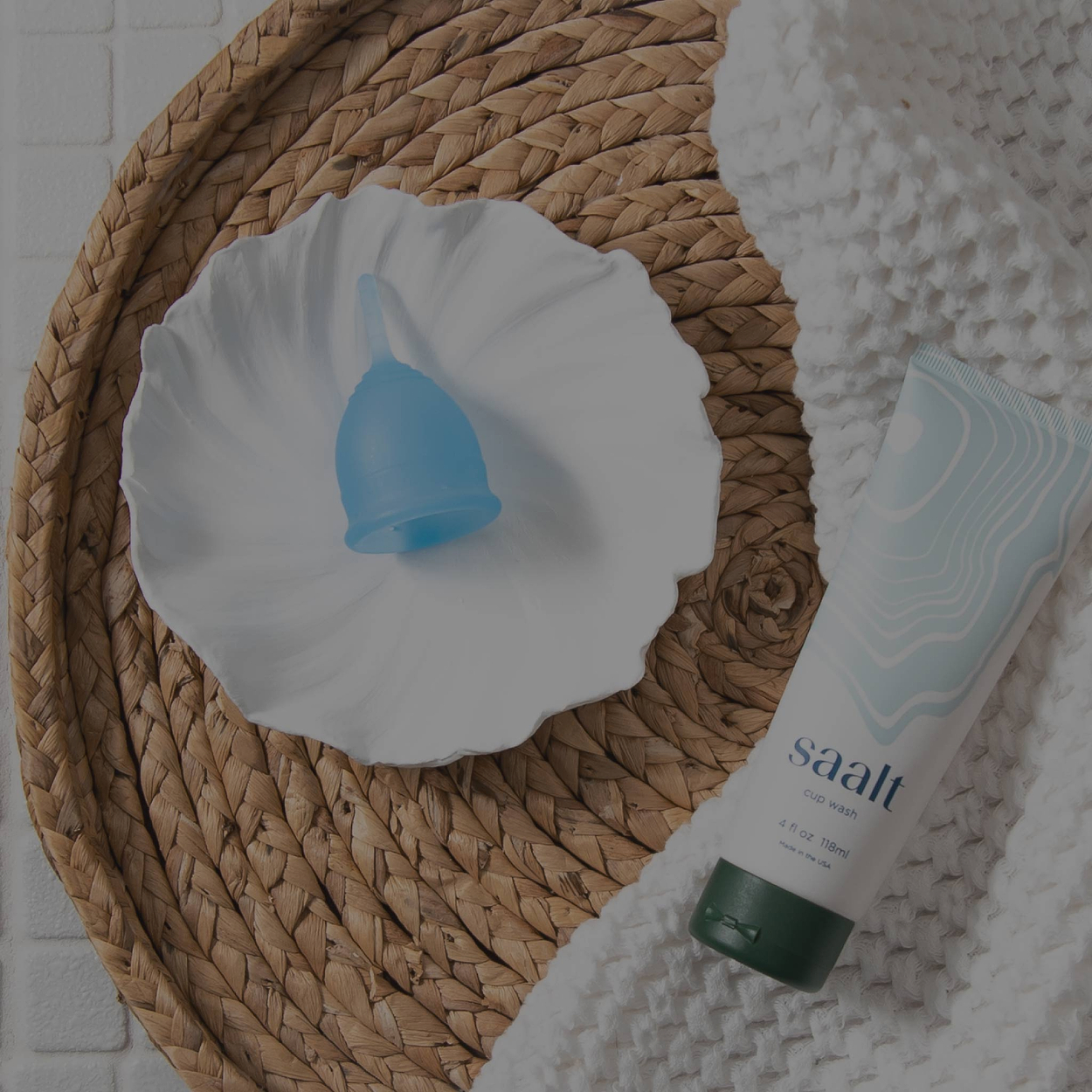 Blue menstrual cup on a white plate next to Saalt cup wash on a woven tray.