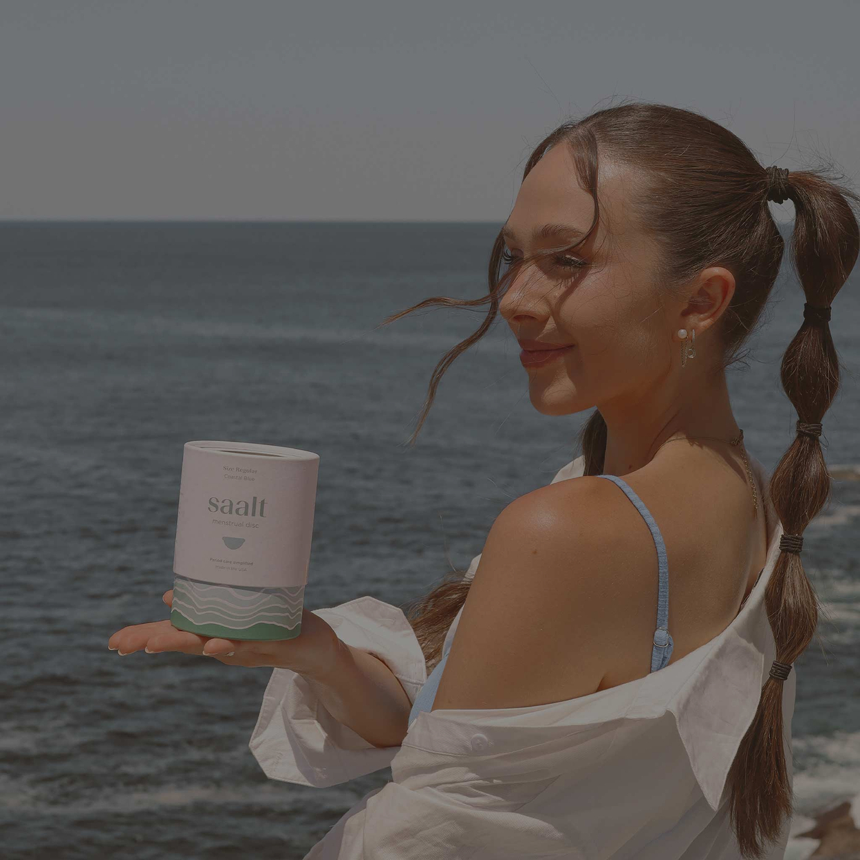 Woman holding a skincare jar by the ocean, wearing a white shirt and blue strap top, hair in a long braided ponytail