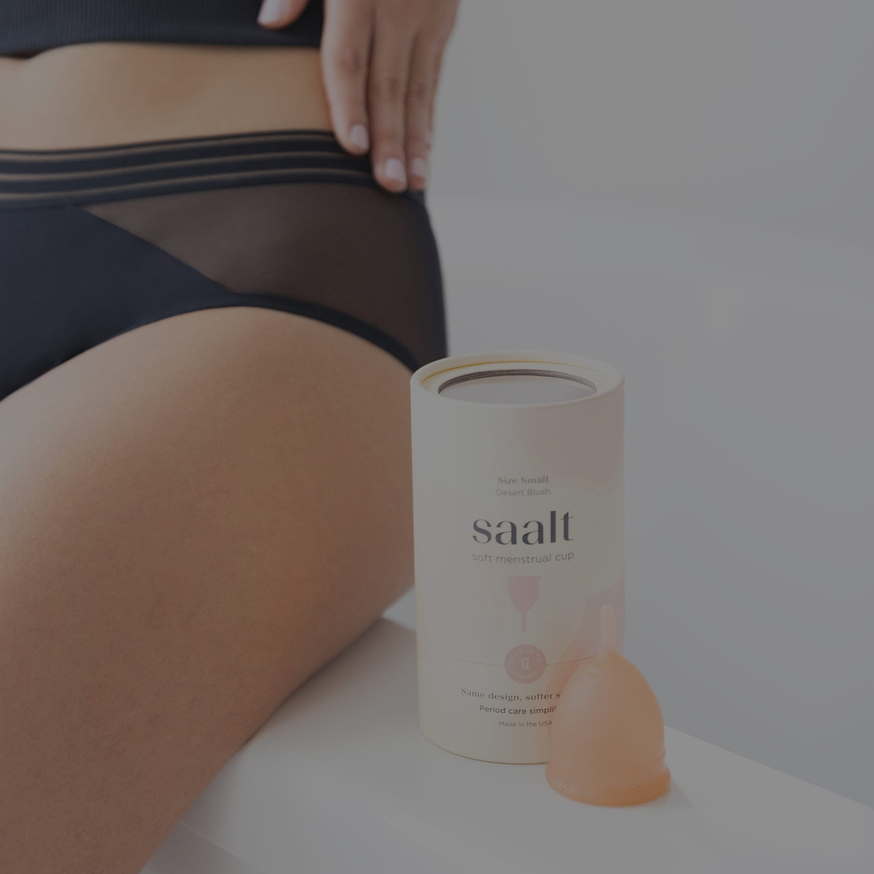 Close-up of a Saalt soft menstrual cup and its cylindrical packaging next to a person in black mesh underwear.