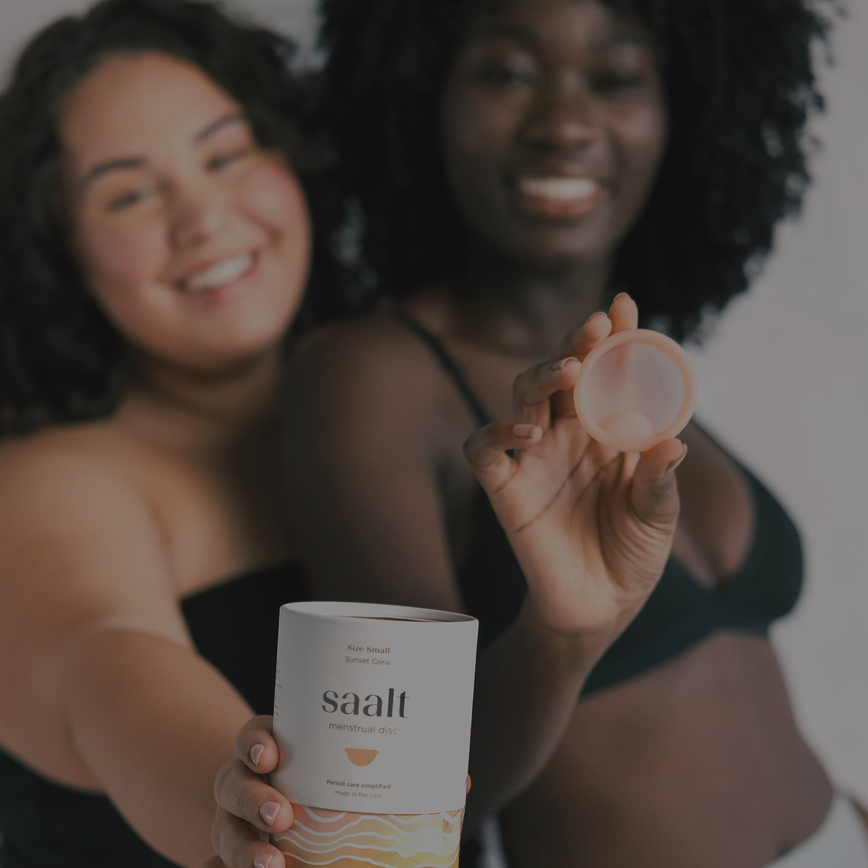 Two women smiling, one holding an open skincare balm and the other a saalt container in the foreground