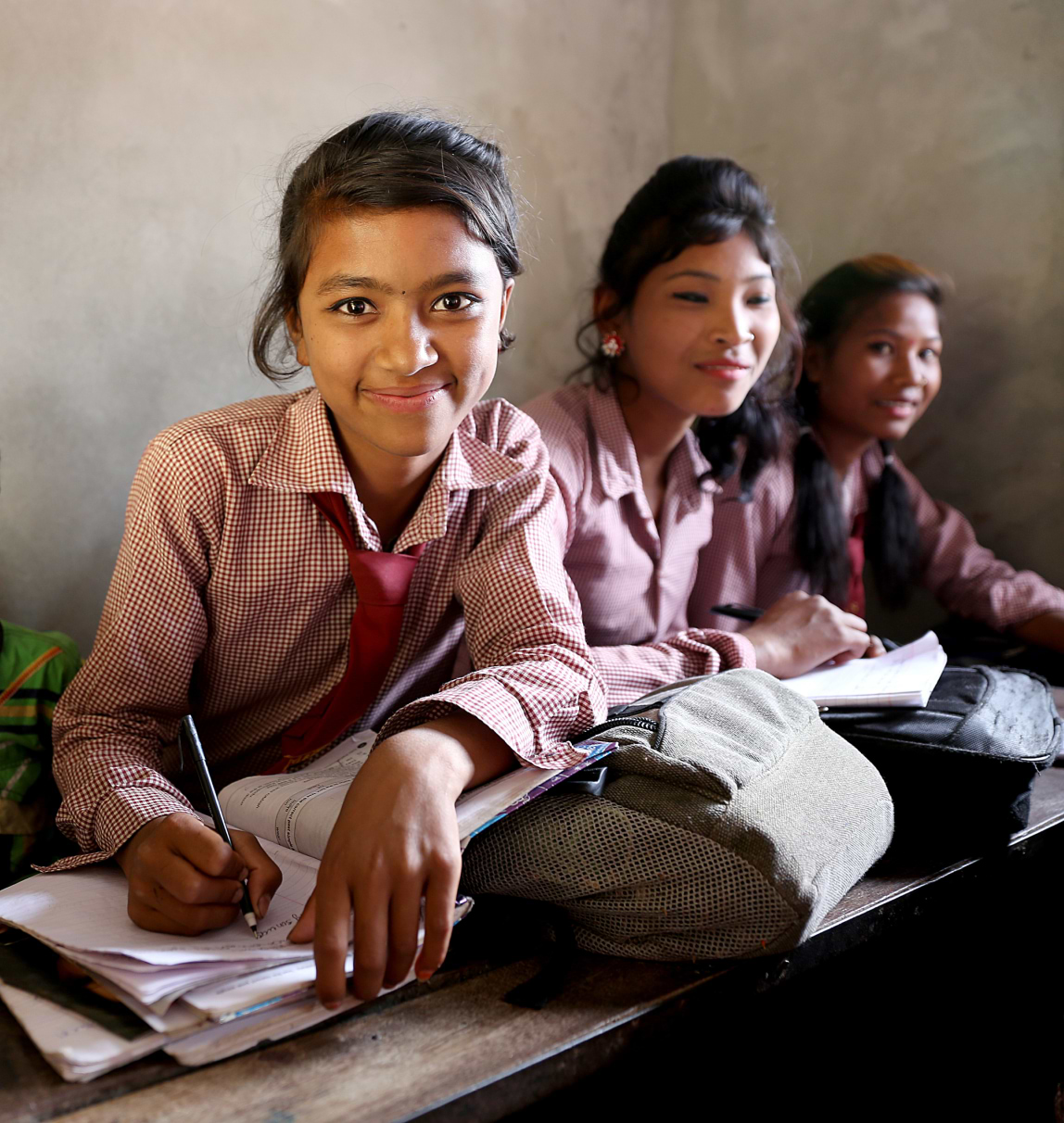 Three smiling schoolgirls in pink uniforms sit at a desk, with the girl in the foreground writing in a notebook.