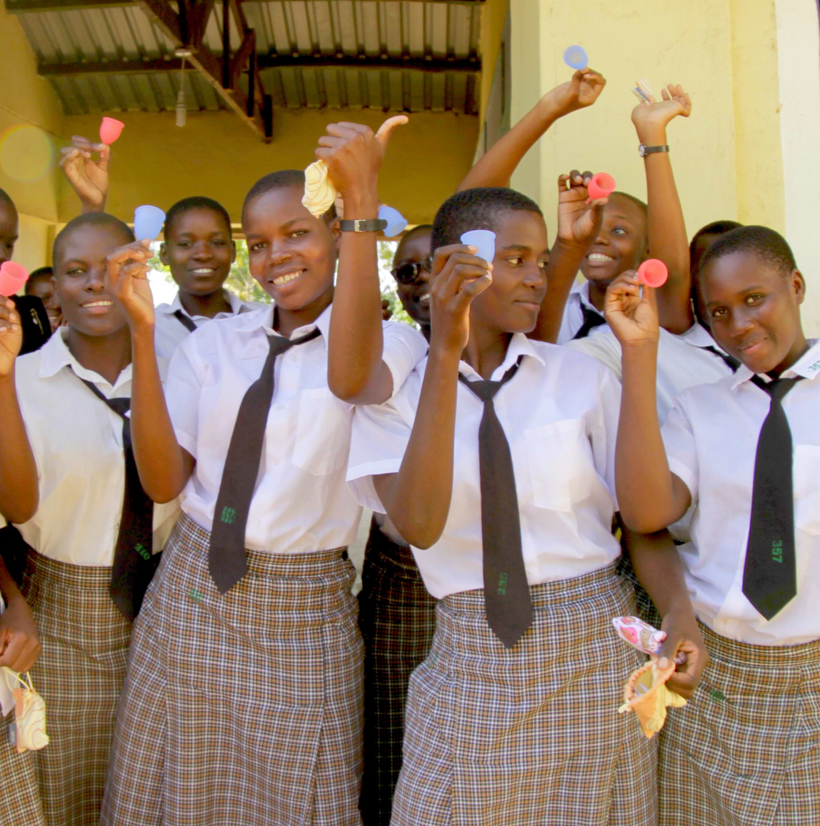 A group of smiling schoolgirls in uniforms holding up various colorful menstrual cups and giving thumbs up.