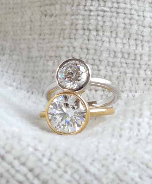 Two bezel set diamond rings, one with a yellow gold band and the other with a white gold band, positioned together on a textured fabric.