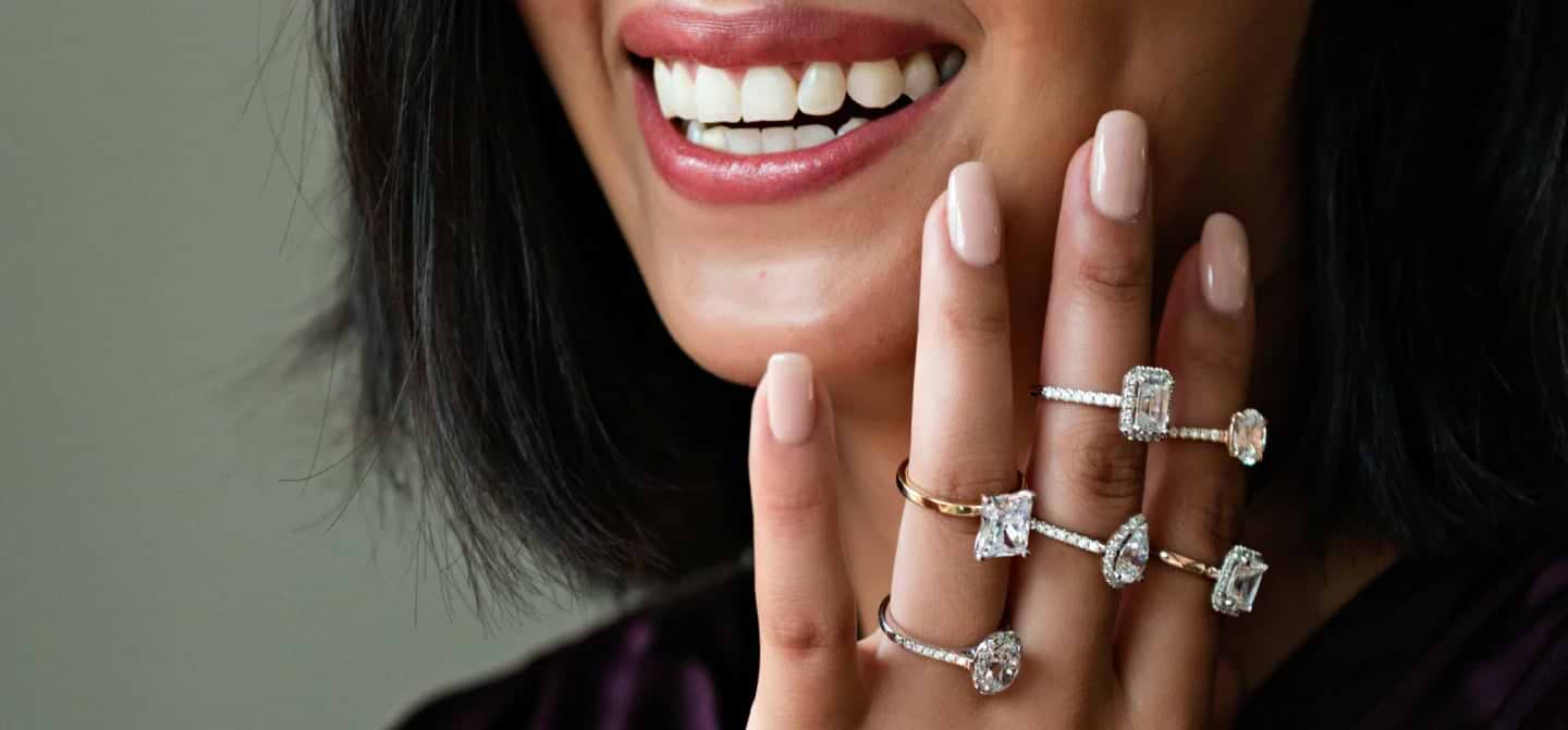 A close up shot of a hand adorned with six large, sparkling diamond rings held up to a woman's face.