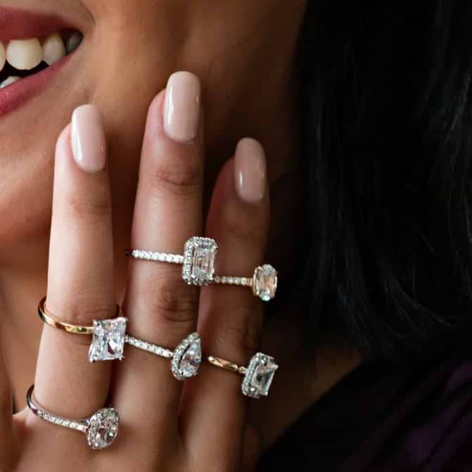 A close up shot of a hand adorned with six large, sparkling diamond rings held up to a woman's face.