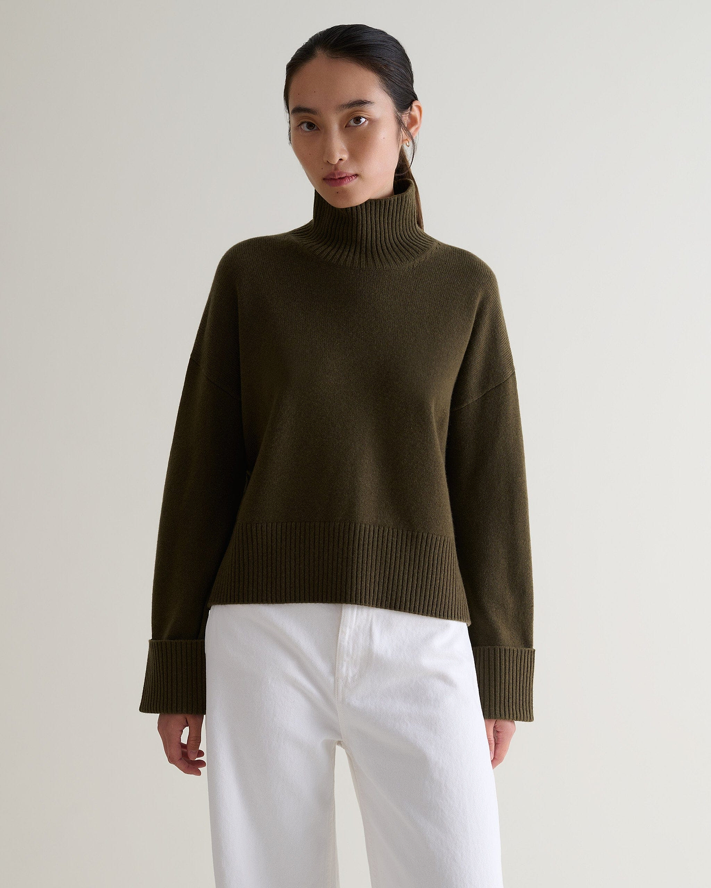 Image 1 of 5 showing woman wearing Rise & Fall's military green cashmere merino high neck jumper with white jeans.