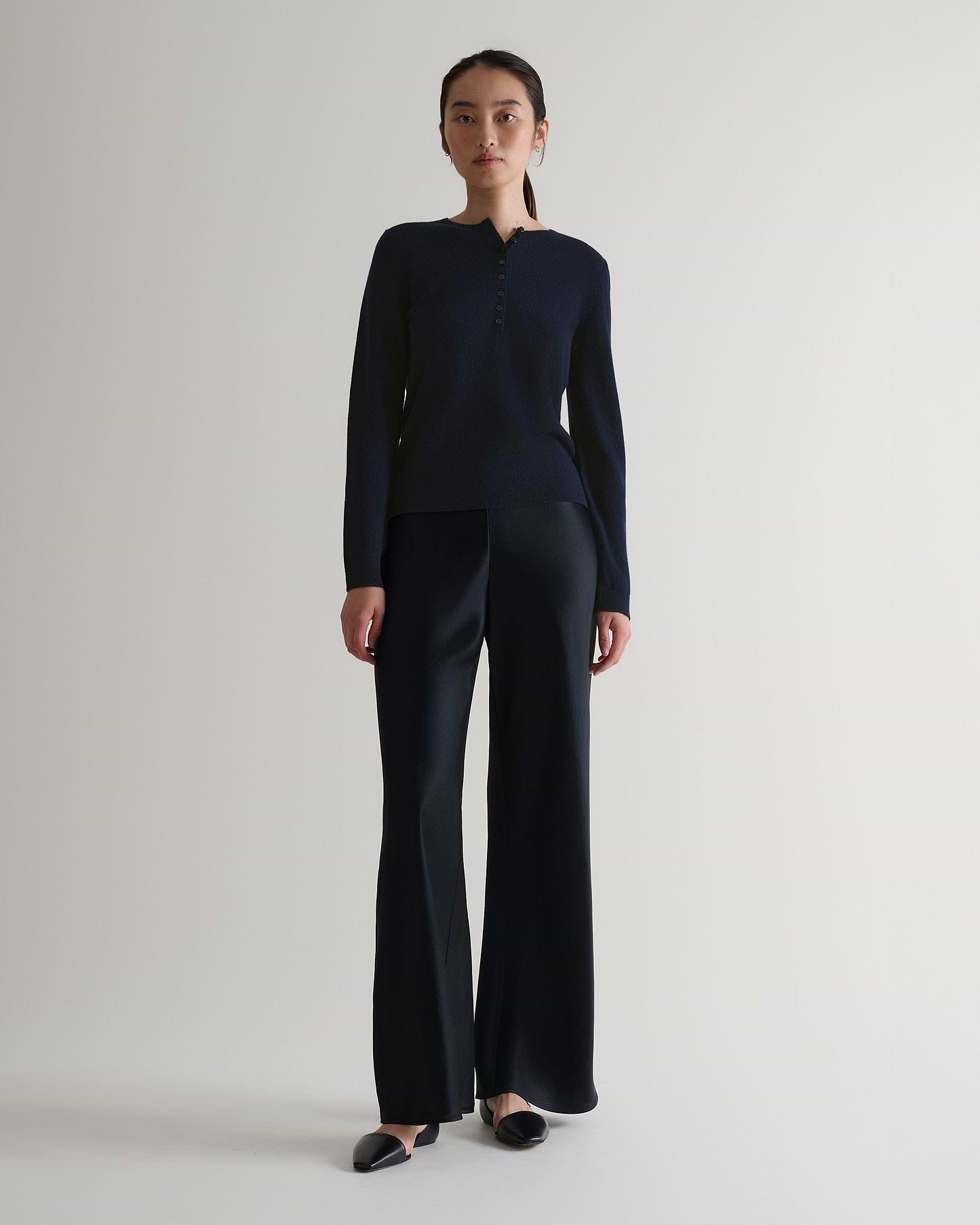 Image 1 of 6 showing a woman wearing Rise & Fall's indigo cashmere merino henley jumper with black silk trousers.