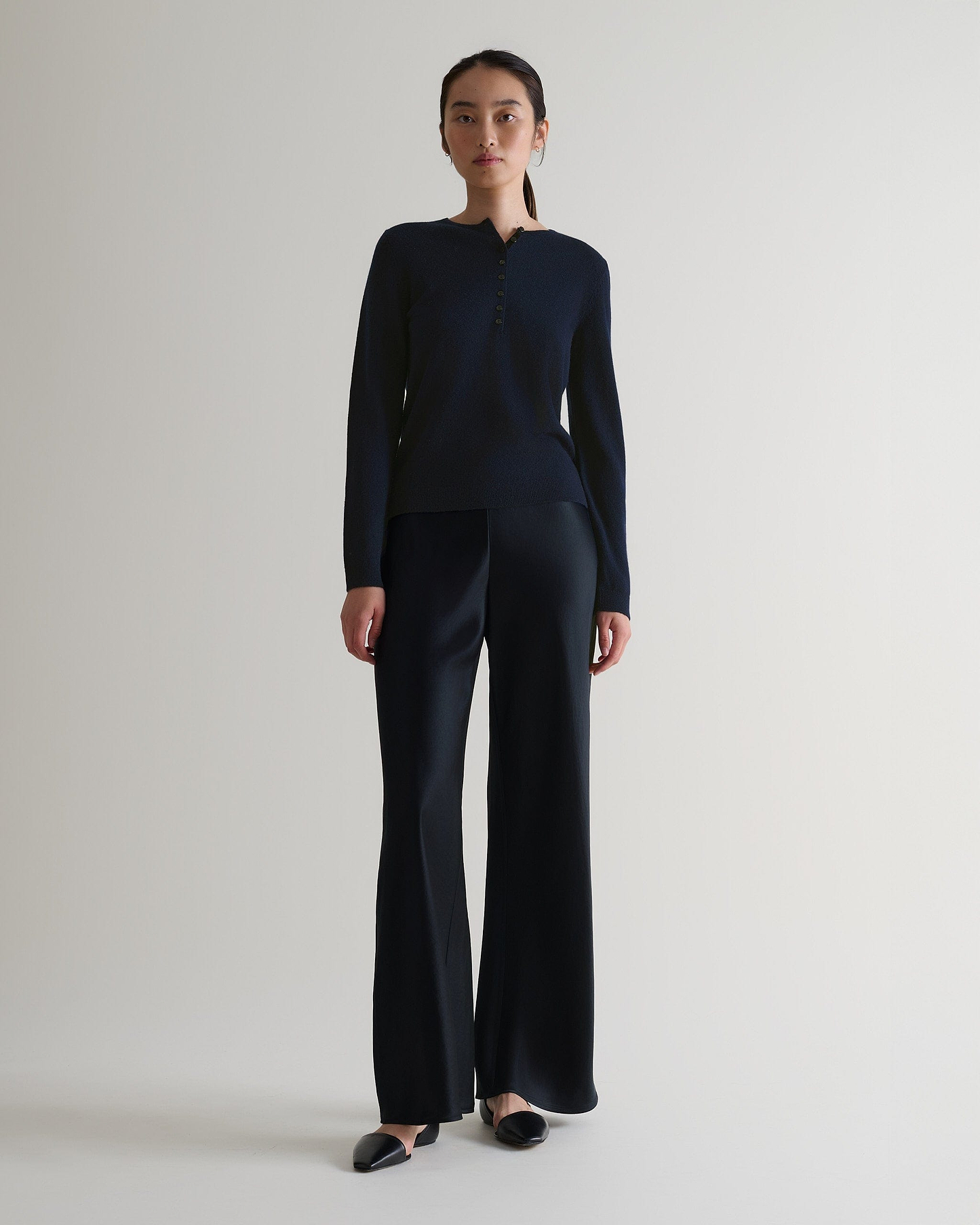 Image 1 of 6 showing a woman wearing Rise & Fall's indigo cashmere merino henley jumper with black silk trousers. 