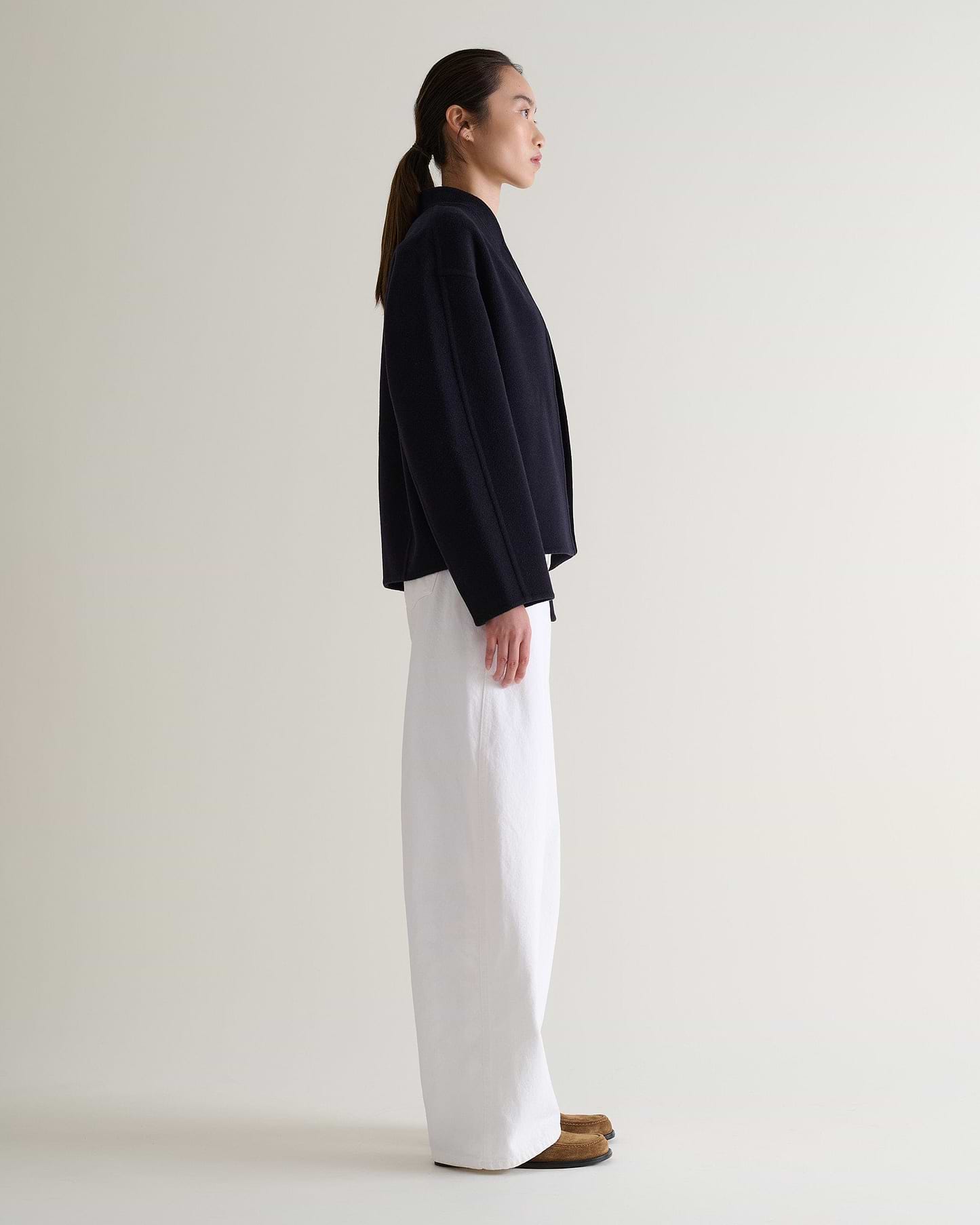 Image 3 of 7 showing side view of woman wearing Rise & Fall's navy cashmere wool double faced scarf jacket with white jeans.