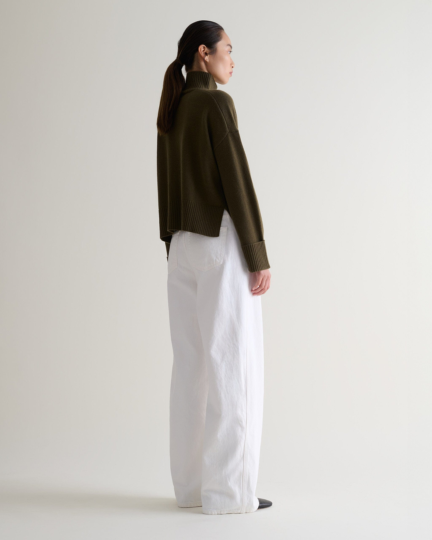 Image 4 of 5 showing back view of a woman wearing Rise & Fall's military green cashmere merino high neck jumper with white jeans. 