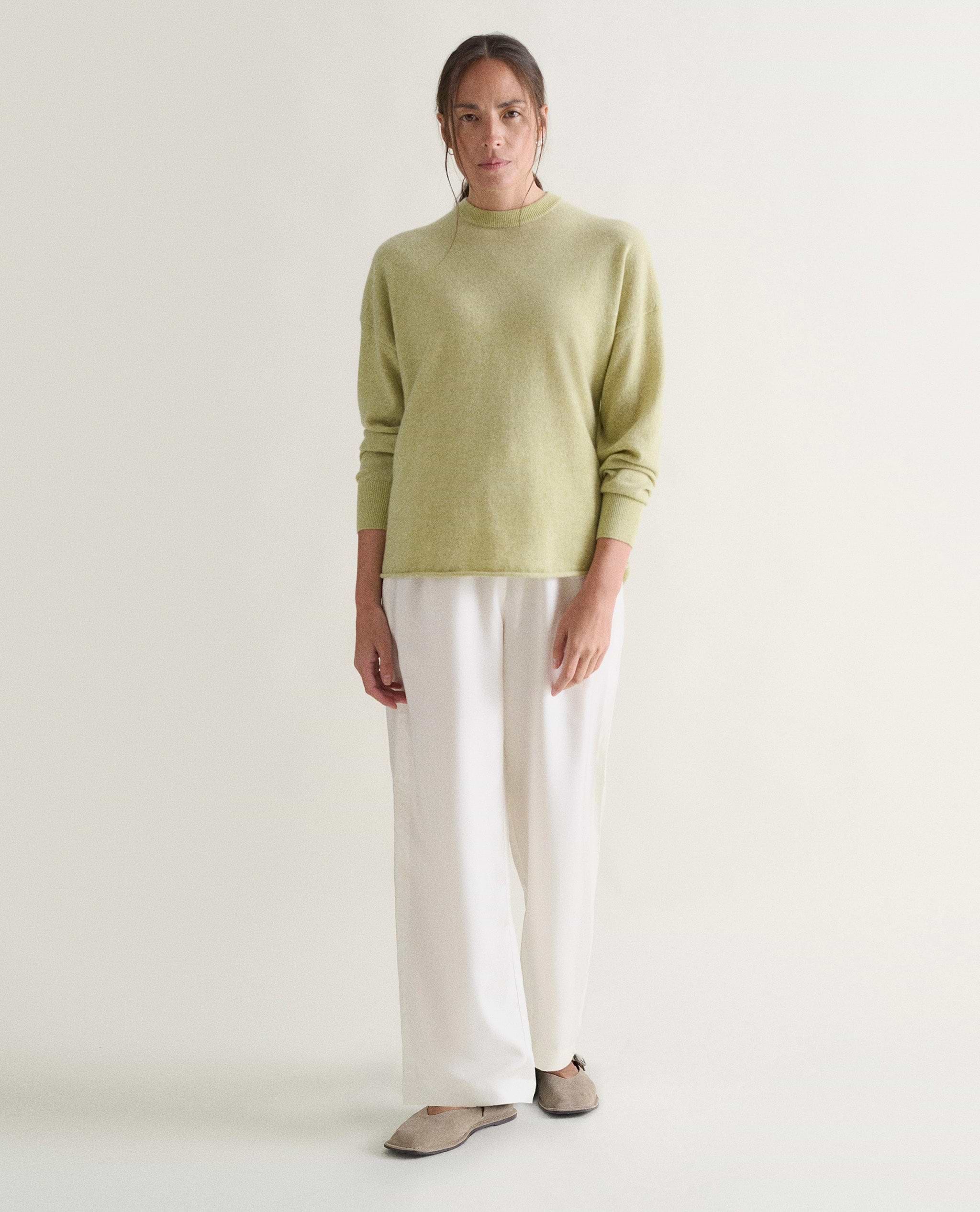 Image 4 of 5 showing woman wearing Rise & Fall's lichen green fine cashmere sweatshirt with white trousers.