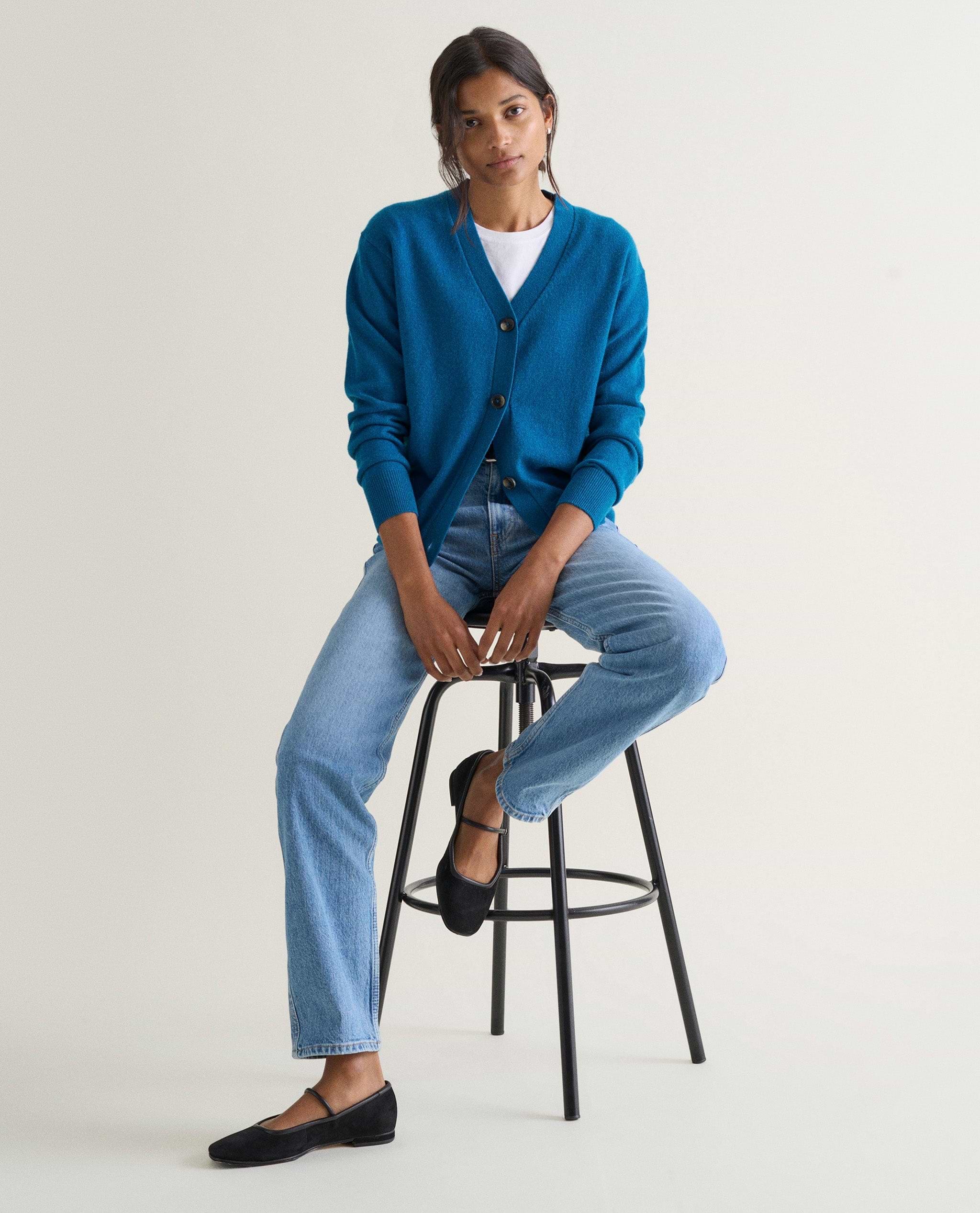 Image 5 of 5 showing a woman sat on a stool wearing Rise & Fall's viridian blue fine cashmere v-neck cardigan with a pair of jeans.
