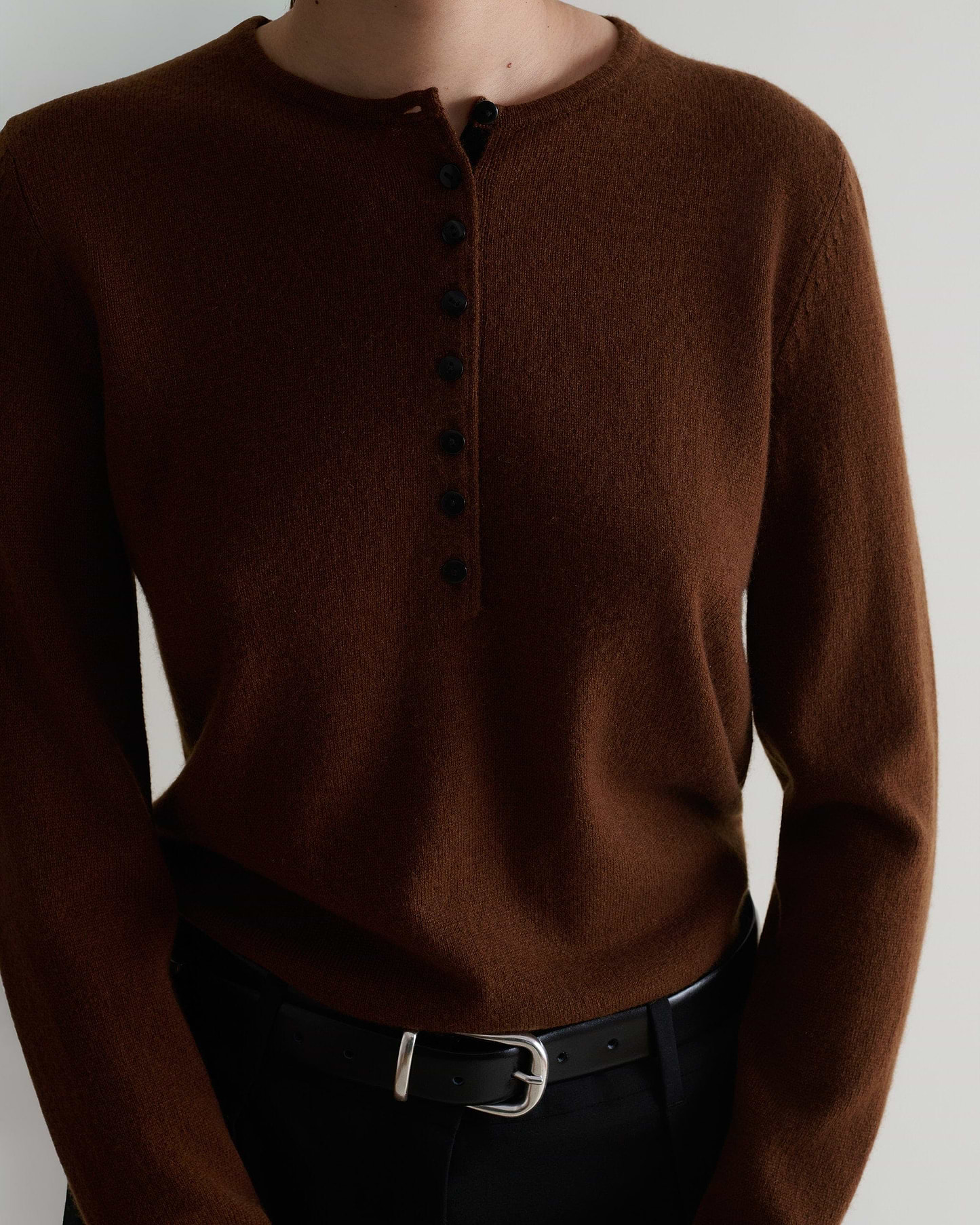 Image 5 of 5 showing a close up of a woman wearing Rise & Fall's brown cashmere merino henley jumper with black trousers and a black belt.