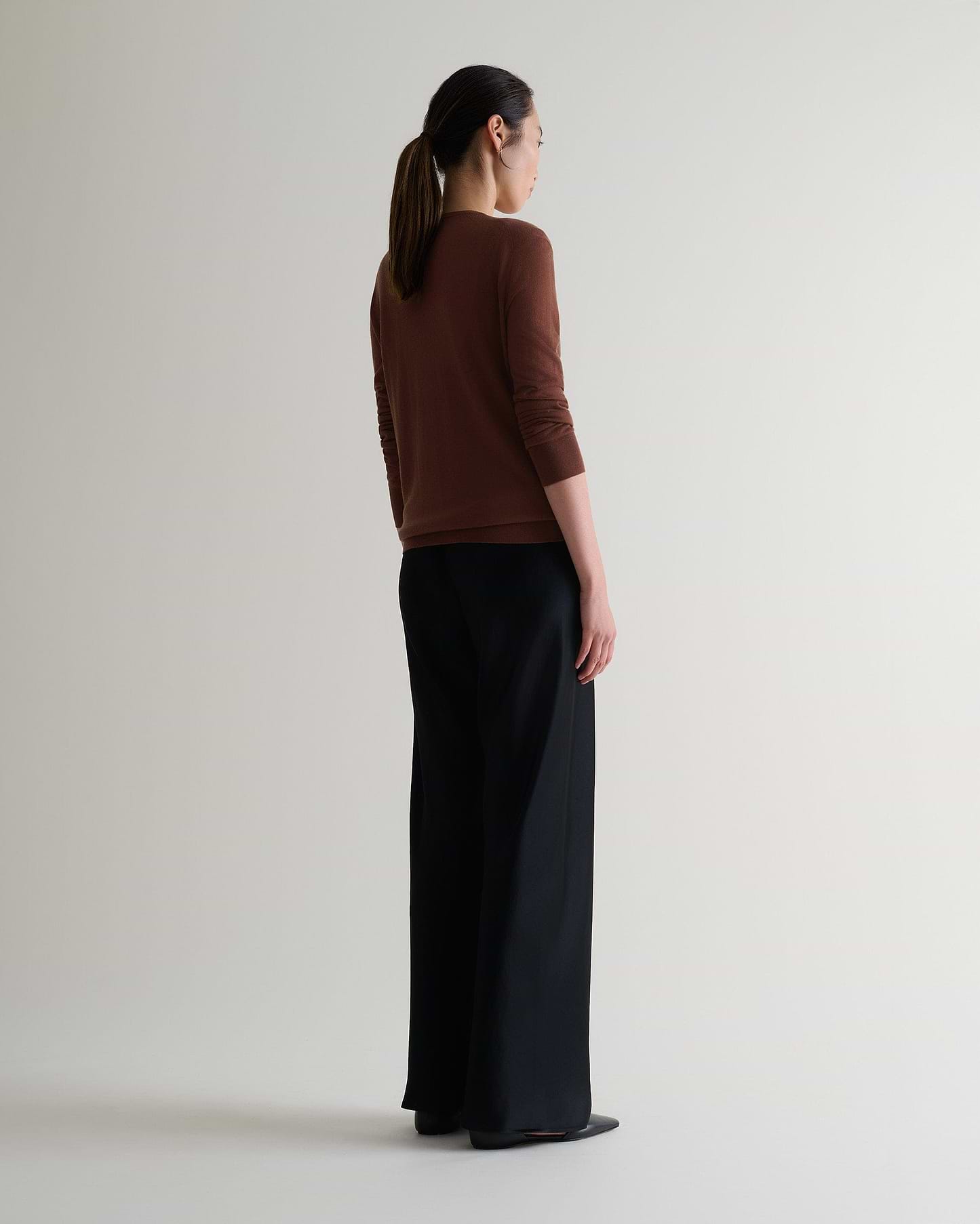 Image 5 of 6 showing full length side view of woman wearing Rise & Fall's brown tencel cashmere double layered crew neck jumper with black trousers.