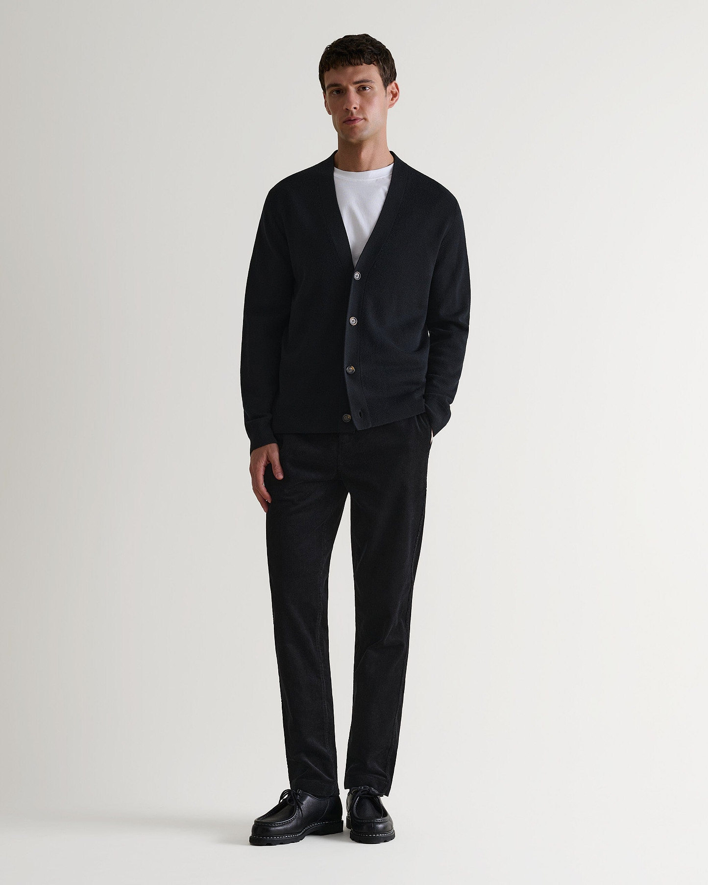 Full length view of man wearing black corduroy trousers with white T-shirt, black cashmere cardigan and black leather shoes.