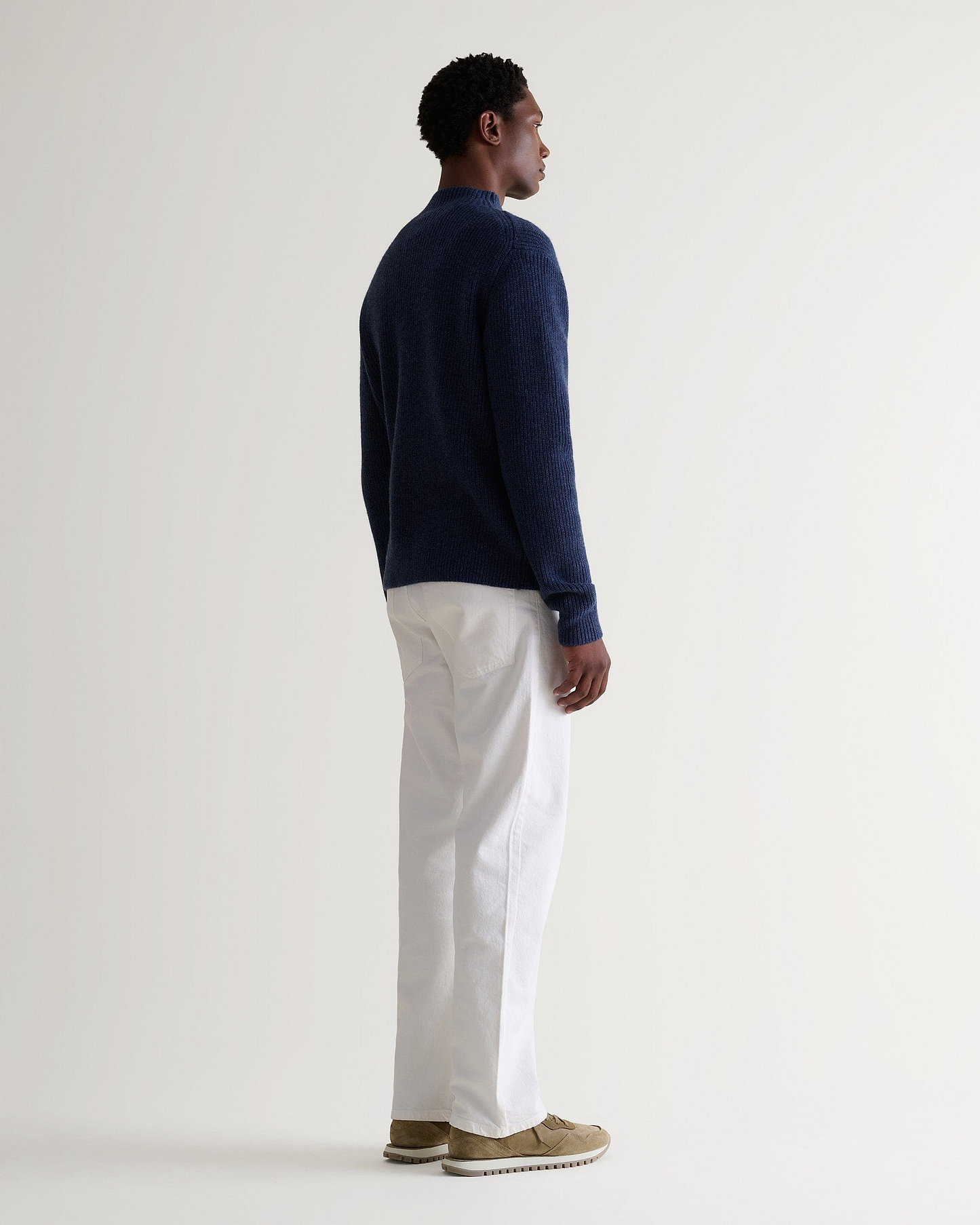 Side view of man wearing Rise & Fall blue cashmere merino stand neck jumper with white trousers and beige suede trainers.