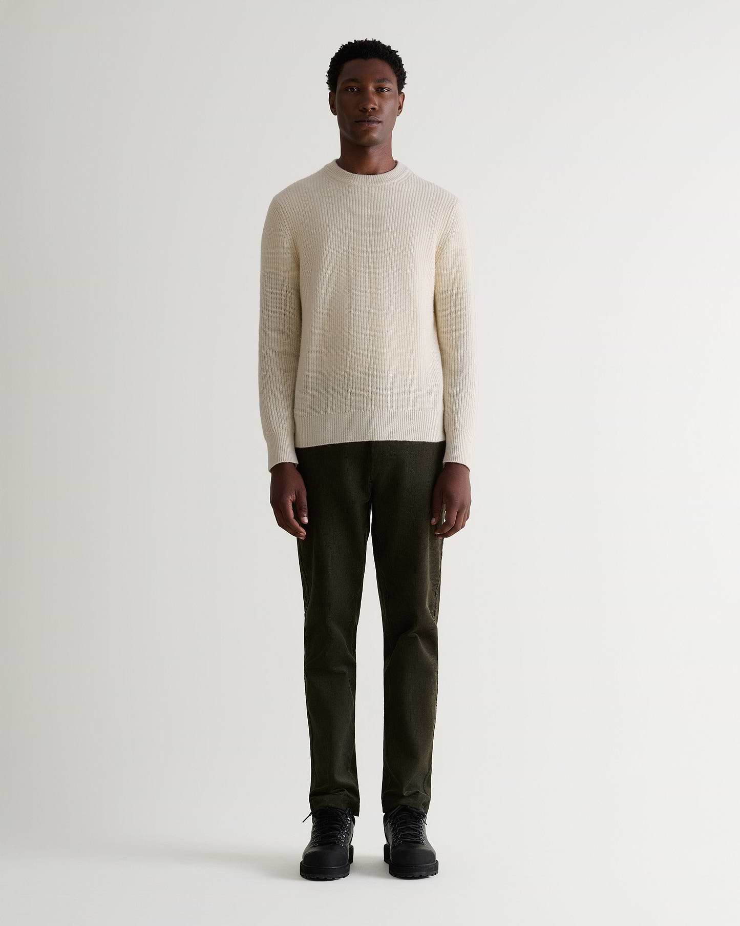 Full length front view of man wearing Rise & Fall's dark green corduroy trousers. Worn with cream jumper and black boots.