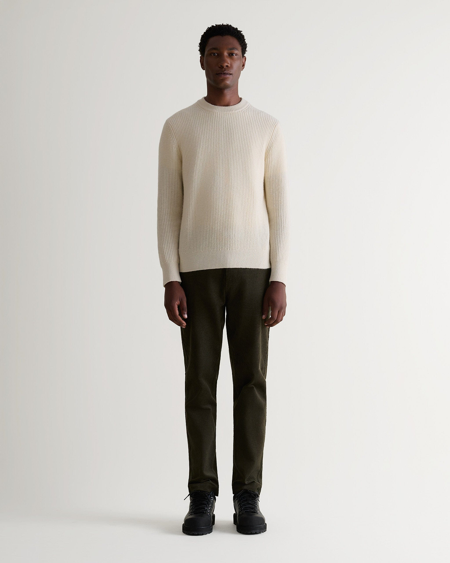 Full length front view of man wearing Rise & Fall's dark green corduroy trousers. Worn with cream jumper and black boots.