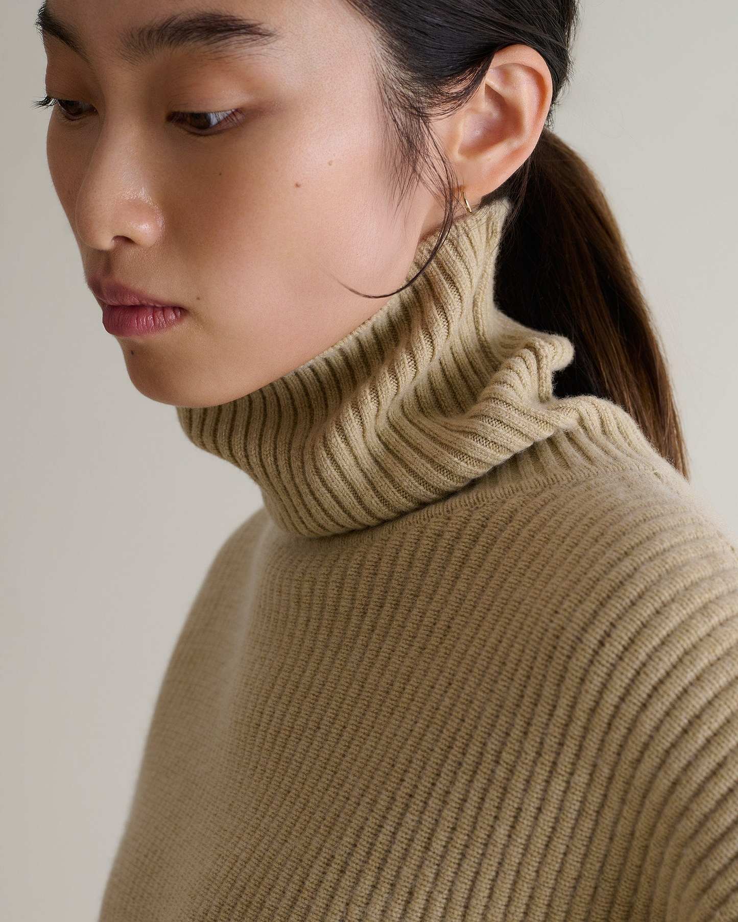 Close up of woman wearing beige cashmere merino Ottoman ribbed jumper with roll neck turned up.