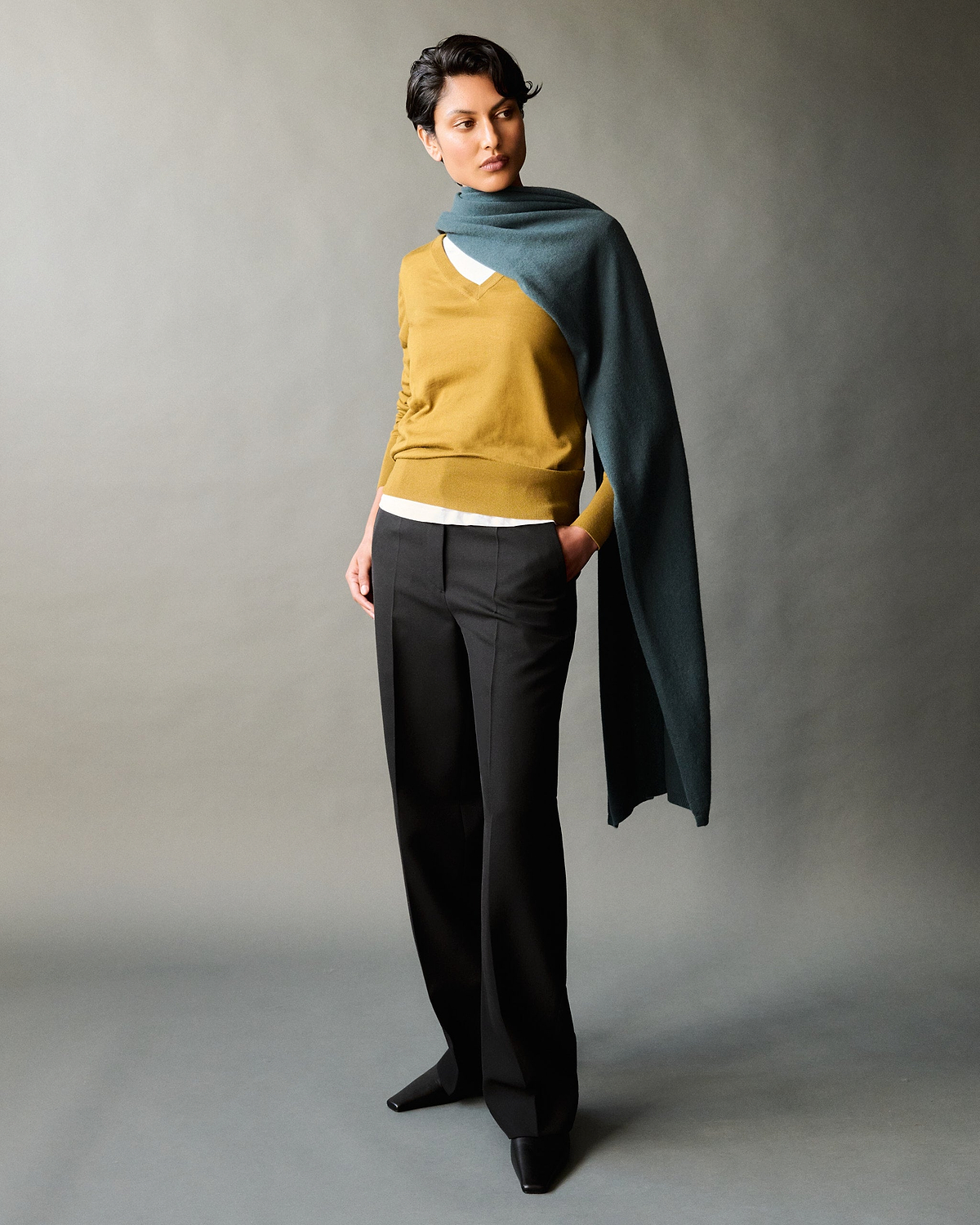 Woman wears Rise & Fall Moonlake Blue Oversized Cashmere Scarf with a v-neck jumper and black trousers.