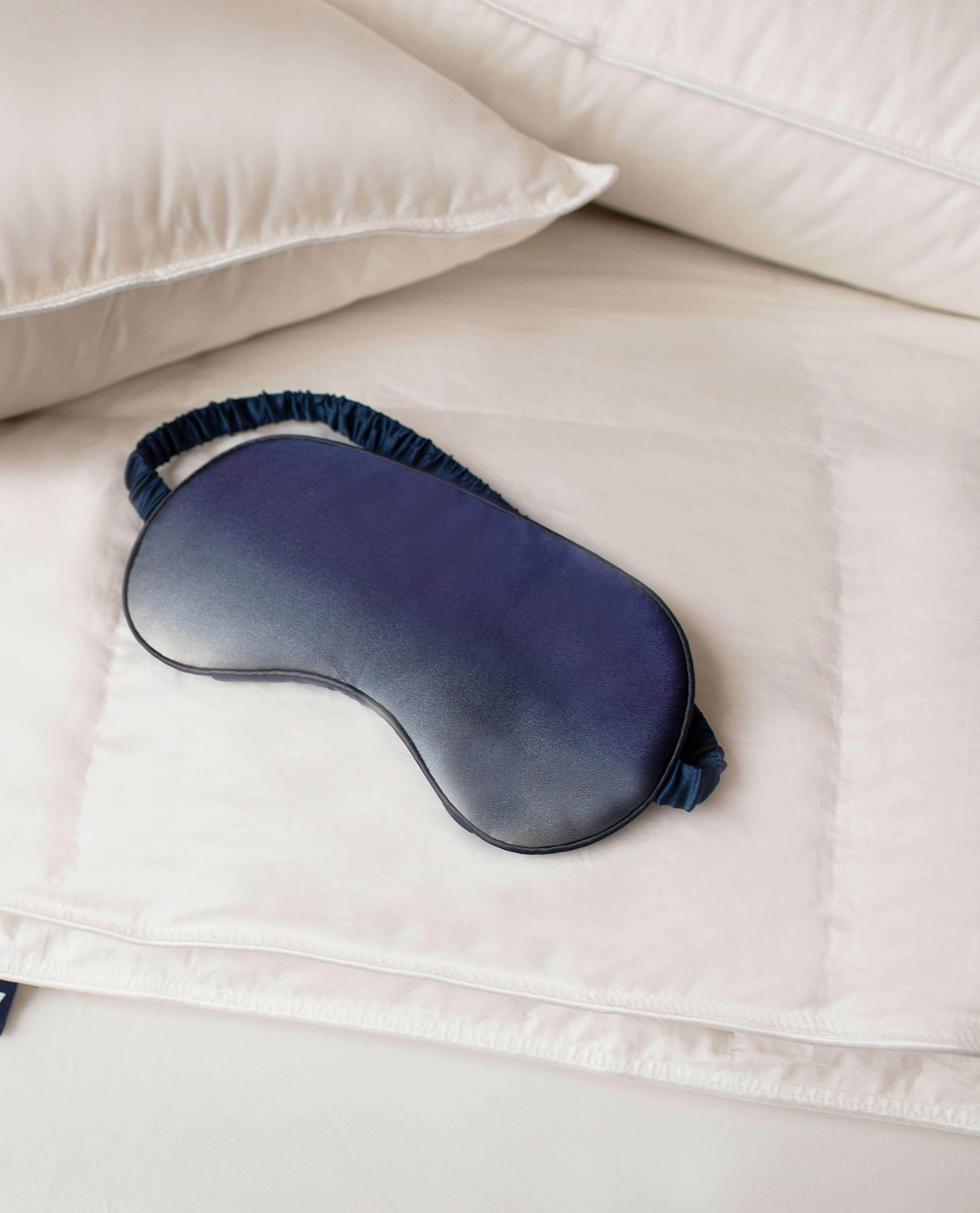 A navy silk sleep mask layering on top of filled bedding.