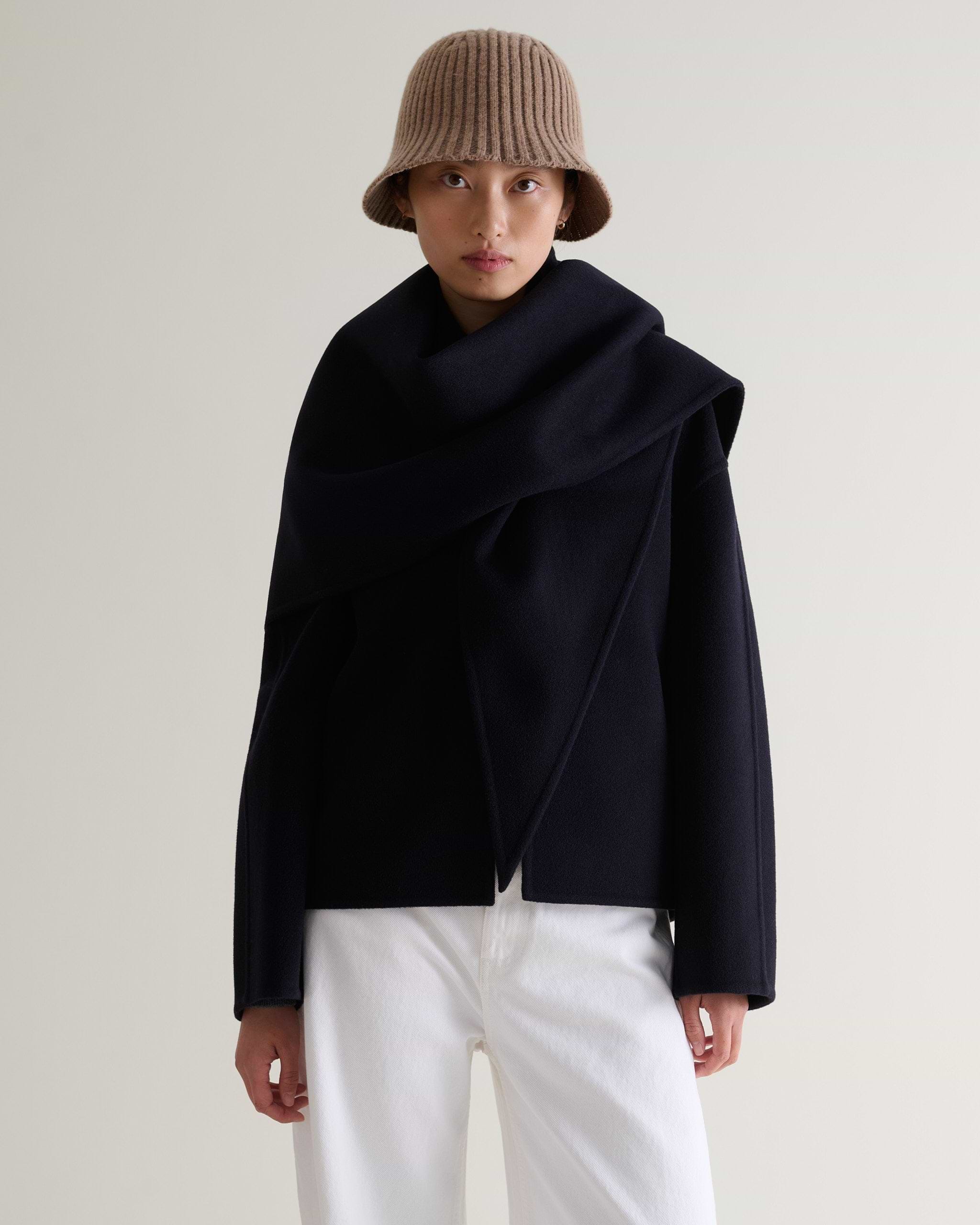 Front view of woman wearing beige merino wool ribbed bucket hat with white trousers and navy cashmere wool scarf jacket.
