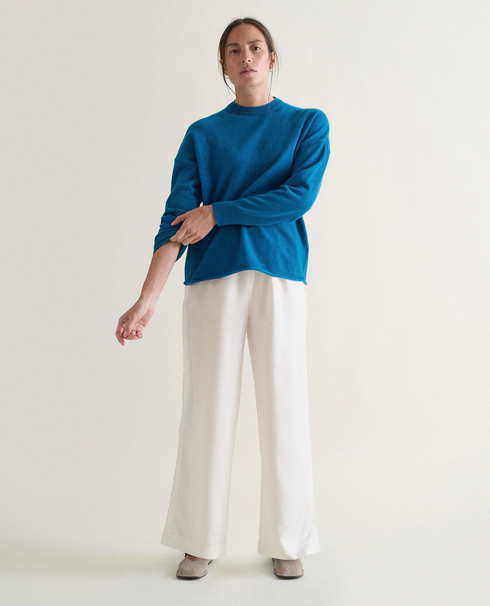 Image 1 of 3 showing woman wearing Rise & Fall's viridian blue fine cashmere sweatshirt with white silk trousers.