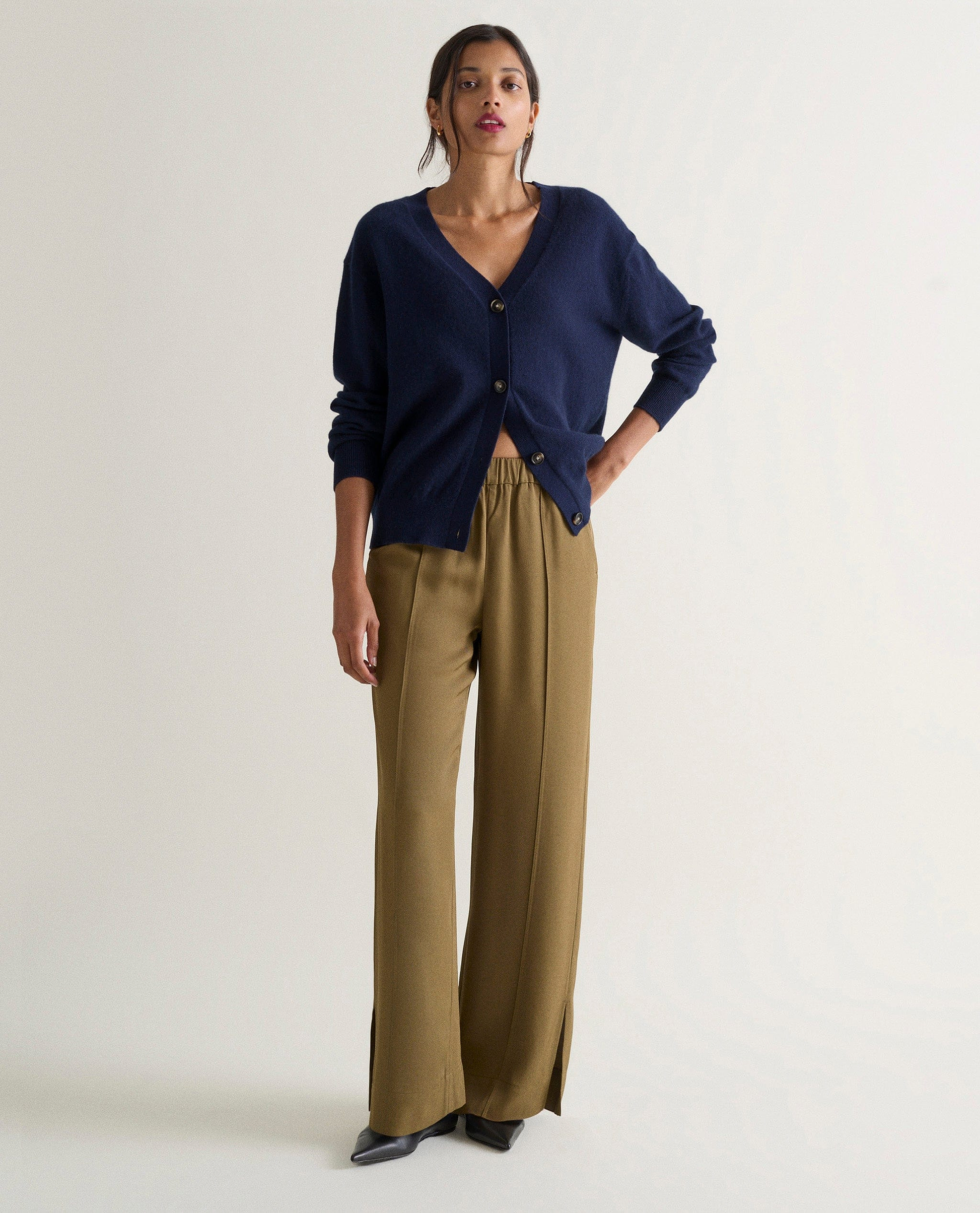 Image 1 of 4 showing woman wearing Rise & Fall's navy fine cashmere v-neck cardigan with olive trousers.