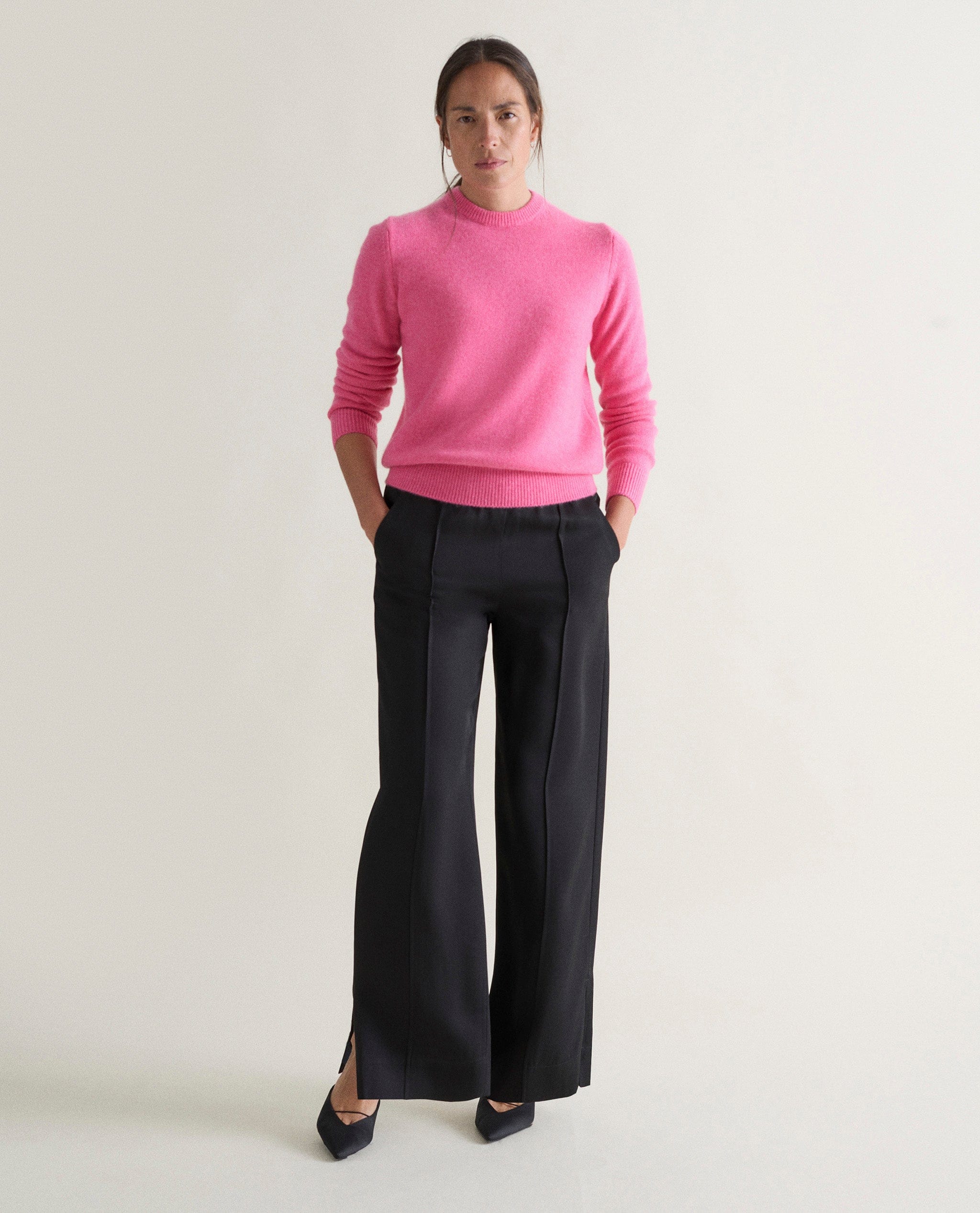 Image 1 of 5 showing woman wearing Rise & Fall's pink mid weight cashmere crew neck jumper with black trousers.