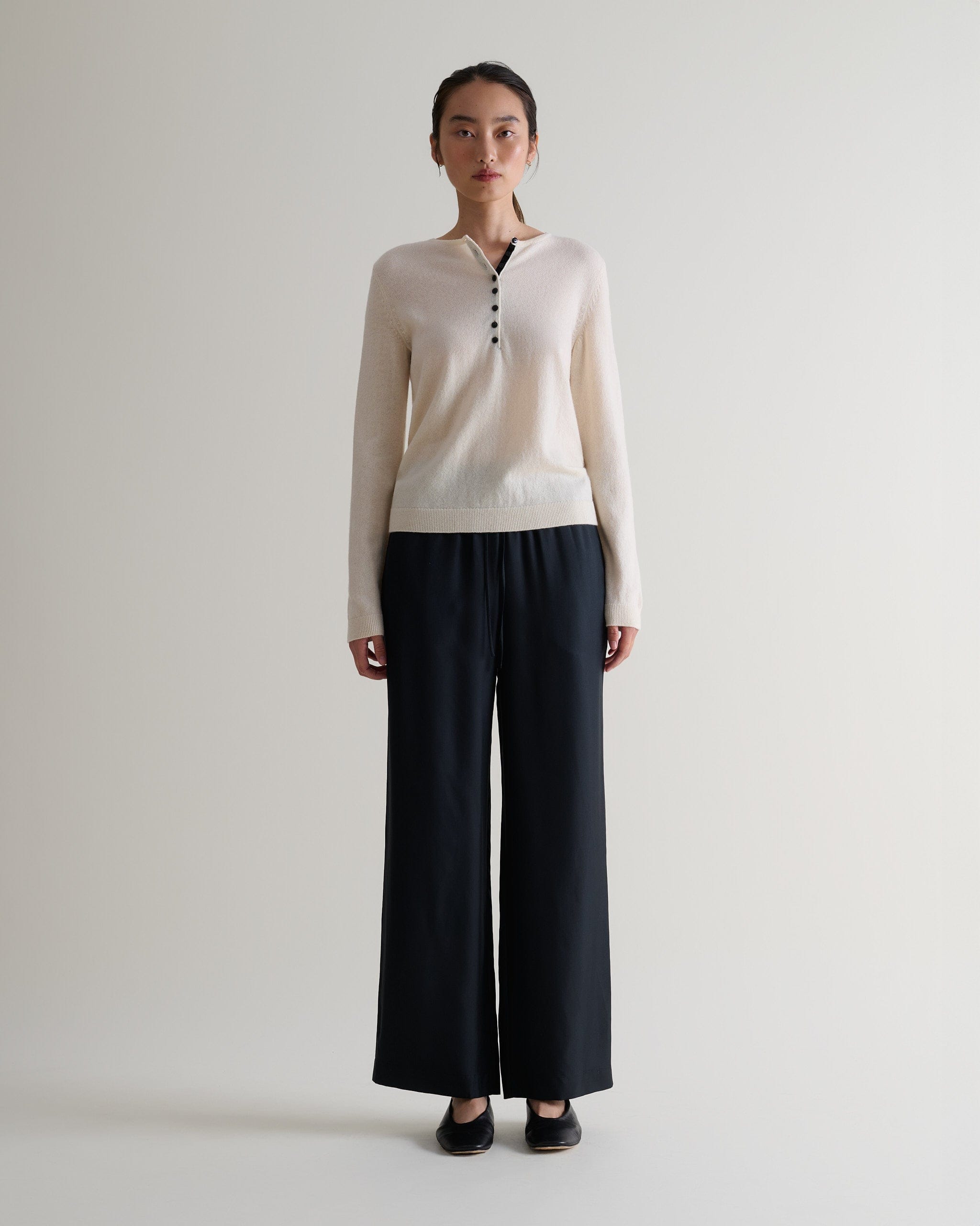 Image 3 of 5 showing a woman wearing Rise & Fall's buttermilk cashmere merino henley jumper with black silk trousers. 